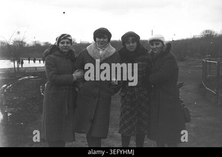 Sloviansk, 1982, in der Nähe eines gefrorenen Teichs. Vier junge Frauen stehen Arm in Arm draußen und posieren warm vor der Kamera an einem kalten Tag. Sie sind in der typischen sowjetischen Wintermode der damaligen Zeit gekleidet und tragen maßgeschneiderte Mäntel, Pelzkragen und Strickmützen. Im Hintergrund können andere Menschen auf einem gefrorenen Gewässer Schlittschuhlaufen sehen. Dieses Archivfoto bewahrt die echte Atmosphäre der Freundschaft, der Damenmode, des sowjetischen Alltags, der historischen Nostalgie und des friedlichen Donbass. Stockfoto