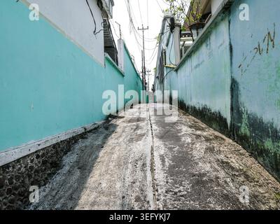 Ein flacher Blick auf eine enge Wohnallee in einer indonesischen Stadt. Der Weg besteht aus verwittertem Asphalt zwischen zwei hohen Wänden, die in einem verblassten gemalt sind Stockfoto