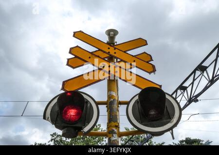 Nahaufnahme eines gelben Bahnübergangs mit einem blinkenden roten Licht vor einem bewölkten Himmel Stockfoto