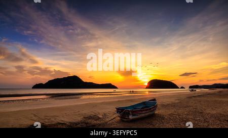 Eine ruhige Küstenszene mit einem verwitterten Holzboot an einem Sandstrand bei Sonnenuntergang, das Themen der Naturreisen und der Einsamkeit am Meer weckt. Stockfoto