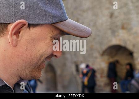 Ein Mann mit grauer Mütze lächelt. Er ist draußen, in der Nähe einer Steinmauer mit sichtbaren Bögen. Im Hintergrund sind Menschen versammelt, einige stehen und Stockfoto