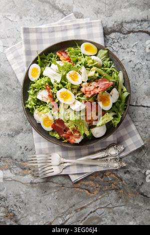 Frischer Salat mit Friseursalat, Mozzarella-Käsebällchen, halbierten gekochten Wachteleiern, knusprigem Speck und Sesamsamen aus der Nähe auf dem Teller auf dem Tisch. Ver Stockfoto