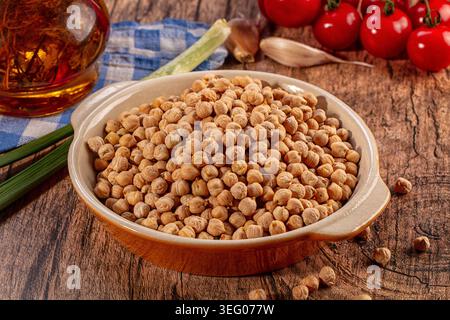 Die Schüssel mit getrockneten Kichererbsen liegt auf einer rustikalen Holzoberfläche. Frische Zutaten wie leuchtende rote Tomaten, grüne Zwiebeln, Knoblauch, reichhaltiges goldenes Öl Stockfoto