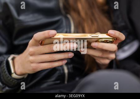 Hände halten das Smartphone für mobiles Gaming. Nahaufnahme eines jungen Menschen, der ein goldenes Smartphone horizontal hält, was auf mobiles Gaming, SMS oder ähnliches hindeutet Stockfoto