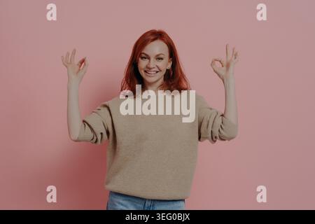 Halblanges Bild einer hübschen Ginger-Frau, die mit beiden Händen OK-Zeichen zeigt, dass alles in Ordnung ist, in lässigem Outfit gekleidet, zufriedenes rothaariges Model, das isoliert über rosa Hintergrund posiert Stockfoto