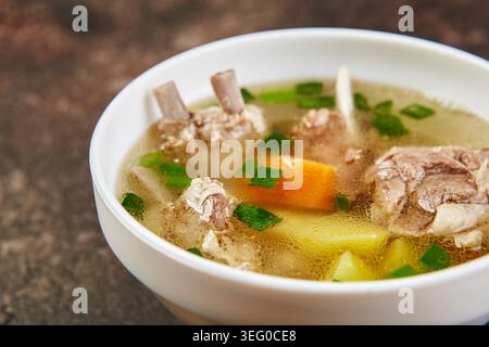 Traditionelle Lammrippsuppe mit Karotten, Kartoffeln und grünen Zwiebeln in einer weißen Schüssel. Klare Brühe, gesunde asiatische Küche, herzhaftes Mittag- oder Abendessen auf dunklem Hintergrund. Stockfoto
