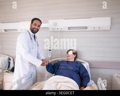 Ein männlicher Arzt aus dem Nahen Osten schüttelt mit einer sich erholenden Patientin, die in einem modernen Krankenhauszimmer eine Augenpflaster trägt, die Hand und vermittelt Vertrauen, mitfühlende Pflege und positive postoperative Erholung. Stockfoto