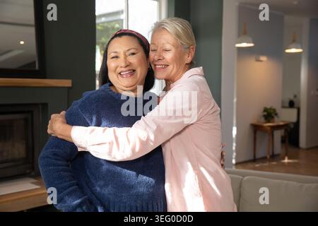Verschiedene ältere Partnerinnen, die sich zu Hause am Fenster umziehen, tragen einen marineblauen Pullover und ein rosafarbenes Hemd. Zuneigung, Gesellschaft, reif, Frau, Wohnzimmer, Sonnenlicht, Couch Stockfoto