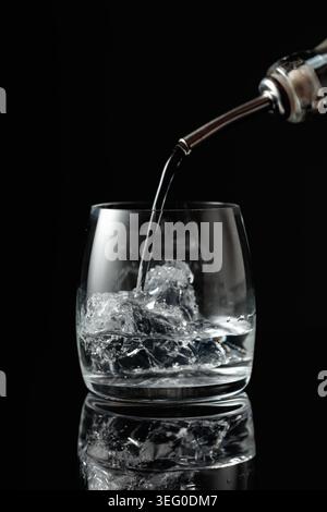 Kristallklarer Wodka oder Gin wird über Eis in ein Glas gegossen. Wodka mit Eis auf schwarzem reflektierendem Hintergrund. Stockfoto
