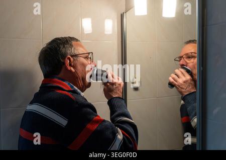 Seniorenmann mit Brille rasiert den Bart mit einem elektrischen Rasierer vor einem Badezimmerspiegel und konzentriert sich dabei auf die tägliche Hygiene Stockfoto