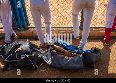 Eine Nahaufnahme mit offenen schwarzen Sporttaschen, gefüllt mit Holzschlägern und Baseballs auf dem roten staubigen Boden eines Sportplatzes in Kuba. Im Hintergrund Stockfoto
