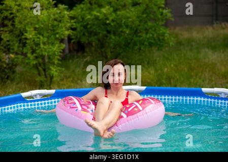 Lächelnde Frau in roten Badeanzügen auf rosa Donut schweben im oberirdischen Pool und genießen einen sonnigen Sommertag in einem grasbewachsenen Hinterhof. Stockfoto