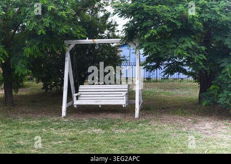 Weiße Gartenschaukel steht zwischen grünen Bäumen im Park. Ein leerer Holzschaukelstuhl bietet Entspannung im Garten während der Sommertage. Klassisches Outdoo Stockfoto