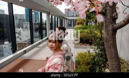 Zwei japanische Frauen im traditionellen Kimono spazieren auf einer modernen Dachterrasse, die von blühenden Sakura-Zweigen, Glasgeländern und einer urbanen Skyline eingerahmt ist Stockfoto