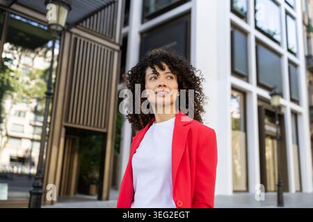 Die hispanische Frau mit lockigem Haar steht selbstbewusst in einer urbanen Umgebung und trägt einen leuchtend roten Blazer. Sie sieht inspiriert aus und ist bereit für neue Opportun Stockfoto