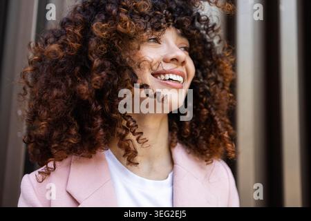 Helle, fröhliche Hispanic-Frau mit lockigem Haar lächelt in einer lässigen Lifestyle-Umgebung warm und zeigt natürliche Schönheit und positive Emotionen auf einer sonnigen Umgebung Stockfoto