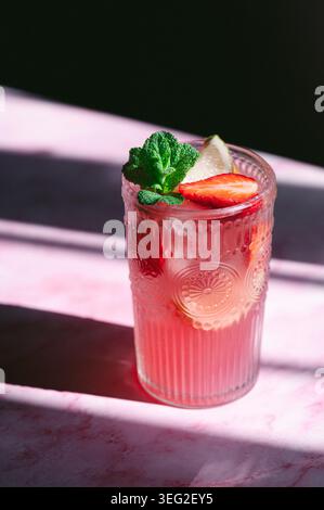Eisige Erdbeerlimonade in einem strukturierten Glas, garniert mit frischer Minze, einem Limettenkeil und Erdbeerscheibe, auf einer rosa Oberfläche mit dramatischer Sonneneinstrahlung Stockfoto