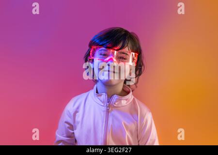 Porträt eines lächelnden Kindes mit leuchtenden LED-Brillen in einem farbenfrohen Neonstudio, das zukünftige Technologie, Innovation, digitalen Lifestyle und pla symbolisiert Stockfoto