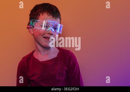 Lächelndes Kind mit futuristischer, intelligenter Brille mit holographischem Leuchten, stehend vor orangem Hintergrund unter Neonlicht, symbolisiert zukünftige Technol Stockfoto