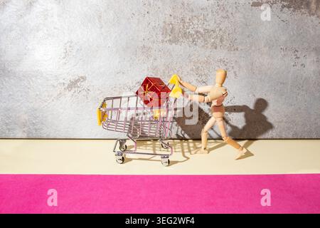 Hölzerne Schaufensterpuppe, die einen Miniatur-Einkaufswagen mit einem rot gewickelten Geschenk-Hintergrund schiebt, mit Stuck-Hintergrund, süßes Geschenk-Konzept Stockfoto