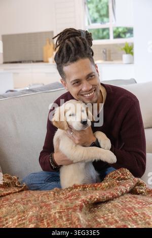 Midadult Afroamerikaner hält goldenen Welpen auf dem Sofa und trägt eine burgunderrote Strickdecke am Fenster Stockfoto