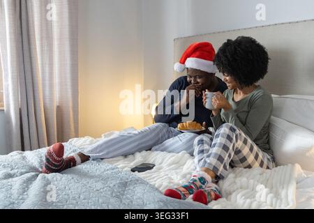 Afroamerikanisches Paar sitzt auf einem Bett im Pyjama und hält Croissant, Tassen und Telefon Stockfoto