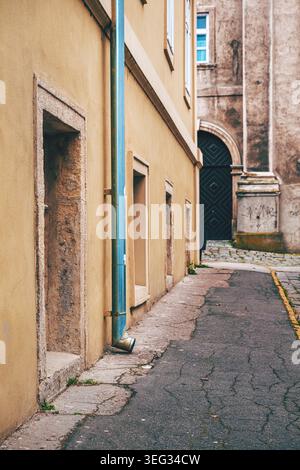 Eine ruhige, enge europäische Gasse, eingerahmt von einem verwitterten gelben Gebäude, blauem downspout und einem bogenförmigen schwarzen Eingang. Strukturierte Stuckwände, Kopfsteinpflaster pavem Stockfoto