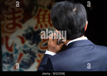 Tokio, Japan. April 2026. Ein japanischer Geschäftsmann oder Büroangestellter in einem klassischen dunkelblauen Nadelstreifenanzug, der auf einem Mobiltelefon spricht, Rückansicht. Der Mann hält das Telefon an seinem Ohr, mit einem hellen Straßenschild mit japanischen Schriftzeichen (Kanji) im unscharfen Hintergrund. Nahaufnahme, Japan. Stockfoto