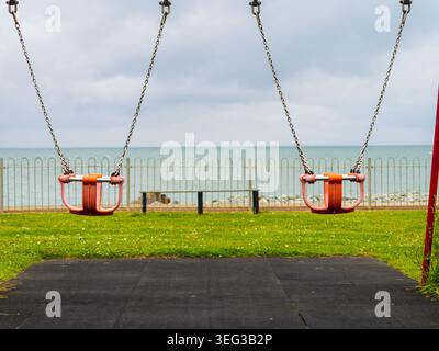 Zwei leere Babyschaukeln mit Blick auf das Meer, die an einem bewölkten Tag in einem Park am Meer in North Wal Gedanken an Kindheit, Spiel und Einsamkeit einladen Stockfoto