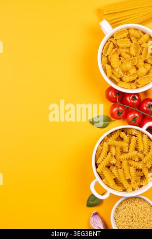 Verschiedene rohe Nudelformen in weißen Schüsseln mit frischen Tomaten, Basilikumblättern, Knoblauch und Spaghetti auf einem hellgelben Hintergrund, die eine lebendige Scul schaffen Stockfoto