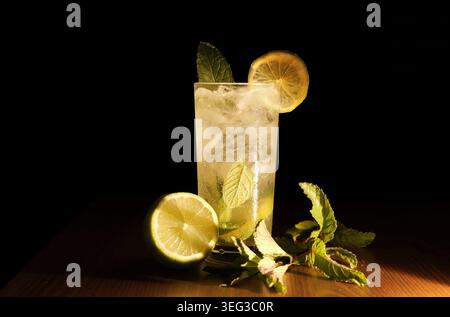Hohes Glas Mojito mit zerstoßenem Eis, verstaubten Minze- und Limettenkeilen, garniert mit Limettenscheibe und Minzzweig auf einem Holztisch vor dunklem Hintergrund Stockfoto