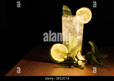 Hohes Glas klassischer Mojito-Cocktail mit Eiswürfeln, lebhaften Minzblättern und einer frischen Limettenscheibe, die auf einer Holzoberfläche vor einem dunklen Hintergrund ruht Stockfoto
