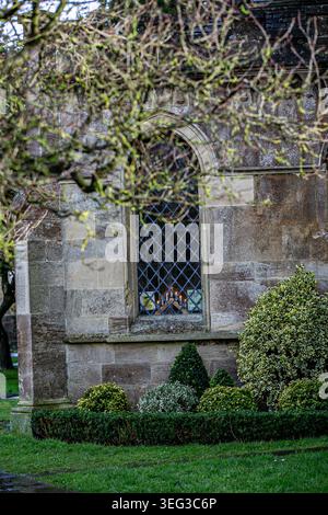 Ein bogenförmiges Fenster im gotischen Stil befindet sich in einer historischen Cotswold-Steinmauer, wahrscheinlich Teil einer Kirche in den Cotswolds. Ein Kerzenständer steht auf dem Fenster Stockfoto