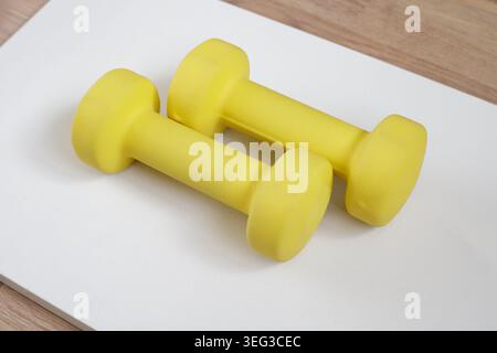 Kleine gelbe Kurzhanteln auf einer hellgrauen Matte oder Vorbereitung für das Heimtraining. Gewichte, Ecke, Fitness, minimalistisch, sonnenbeleuchtet, Boden, wiederverwendbar. Sportausrüstung Stockfoto