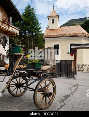 Matrei in Osttirol, Österreich - 5. Juli 2021: Hölzerner Pferdewagen mit alten Rädern und Blumentopf steht neben einer kleinen gelben Almkapelle mit Glocke Stockfoto
