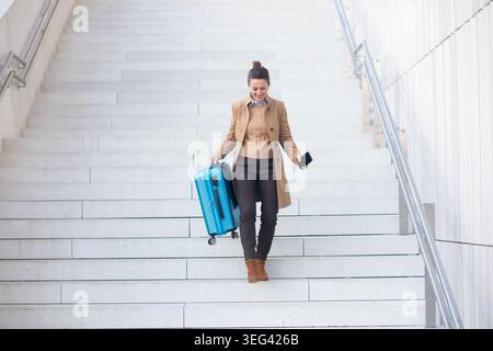 Eine lächelnde Frau in einem braunen Mantel überprüft ihr Handy, während sie an einem Bahnhof mit einem blauen Rollkoffer eine moderne Treppe hinabsteigt. Stockfoto