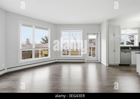 Helles, leeres Zimmer mit Hartholzfußboden und weißen Wänden. Große Fenster lassen Tageslicht einströmen und bieten einen Blick auf den Außenbereich. Küchenbereich sichtbar mit moderner ap Stockfoto