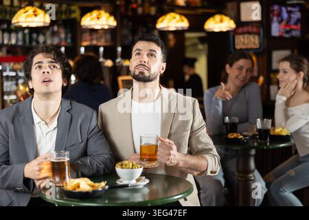 Zwei fröhliche junge Männer, Freunde, die sich unterhalten, Spiele im Fernsehen anschauen und Bier am Tisch im Pub trinken Stockfoto