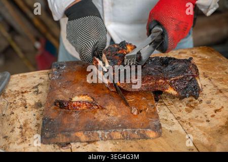 Der Koch schneidet die Brust auf einem gewürzten Holzbrett, trägt schwarze und rote Schutzhandschuhe und verwendet Messer und Zange, um rauchige Speisen zuzubereiten Stockfoto