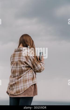 Frauen-Smartphone-Himmel fängt den bewölkten Horizont ein, während sie an einem bewölkten Tag eine karierte Hemdjacke tragen Stockfoto