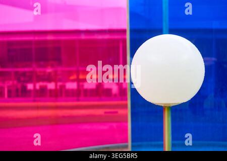 Eine Nahaufnahme einer weißen sphärischen Lampe oder Leuchte, die vor lebendigen farbigen Glasscheiben platziert ist, mit hellem magentafarbenem Rosa links und Stockfoto