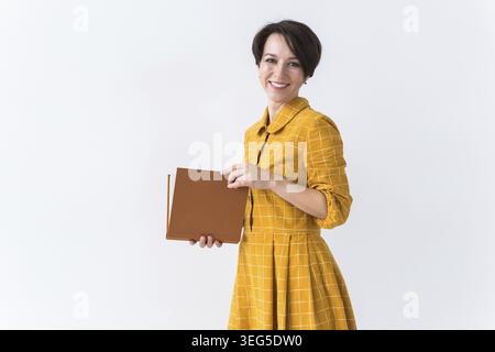 Porträt eines schönen brünetten Mädchens in einem gelben Retro-Kleid, das ein Buch auf weißem Hintergrund liest Stockfoto