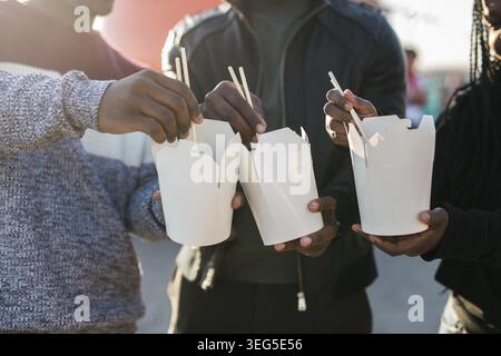 Drei afroamerikanische Freunde genießen an einem sonnigen Tag asiatisches Streetfood im Freien, mit Essstäbchen und mit zwanglosen Momenten in der Nähe eines leuchtenden Orange Fo Stockfoto