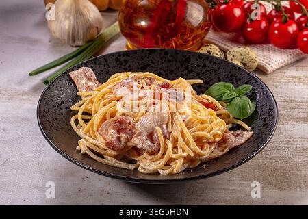 Creme Spaghetti Carbonara in schwarzer Schüssel, garniert mit frischem Basilikum. Knusprige Speckstücke, reichhaltige Sauce. Hintergrund zeigt Knoblauch, Kirschtomate Stockfoto