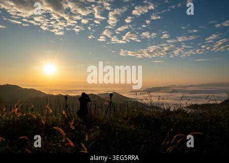 Eine Silhouette eines Fotografen mit Stativ dokumentiert einen dramatischen Sonnenaufgang über einer Landschaft, die im Morgennebel bedeckt ist und ein Abenteuer darstellt. Stockfoto
