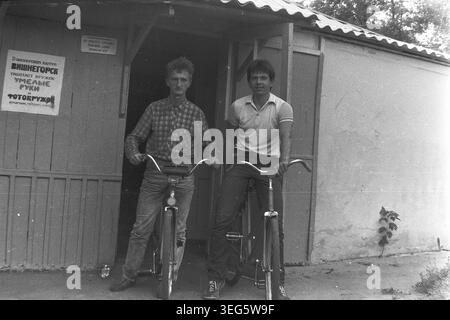 Slawjansk, Ukrainische SSR, 1983. Vintage-Schwarzweiß-Fotografie aus dem Pionierlager Vishegorsk in der Gegend von Slavkurort. Das Bild zeigt zwei junge Mitarbeiter, darunter Dmitri Michailowitsch Kravelis, die selbstbewusst auf klassischen Fahrrädern sitzen. Sie posieren vor einem Holzgebäude mit einem Schild für den Fotoclub. Dieses authentische Archivbild fängt die Atmosphäre der sowjetischen Jugendkultur, des alltäglichen Lebensstils und der Sommererholung ein. Ein nostalgischer historischer Einblick in die Vergangenheit, der einen friedlichen Donbass darstellt. Stockfoto