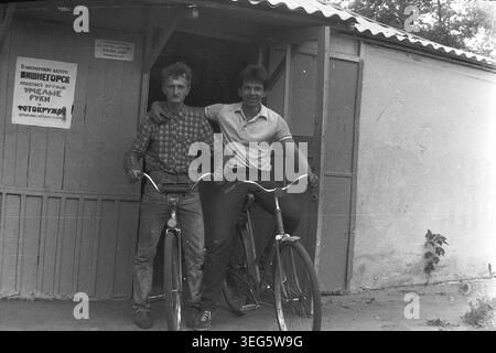 Slawjansk, Ukrainische SSR, 1983. Vintage-Schwarzweiß-Fotografie aus dem Pionierlager Vishegorsk in der Gegend von Slavkurort. Das Bild zeigt zwei junge Mitarbeiter, darunter Dmitri Michailowitsch Kravelis, die selbstbewusst auf klassischen Fahrrädern sitzen. Sie posieren vor einem Holzgebäude mit einem Schild für den Fotoclub. Dieses authentische Archivbild fängt die Atmosphäre der sowjetischen Jugendkultur, des alltäglichen Lebensstils und der Sommererholung ein. Ein nostalgischer historischer Einblick in die Vergangenheit, der einen friedlichen Donbass darstellt. Stockfoto