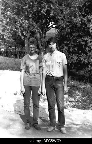 Slawjansk, Ukrainische SSR, 1983. Vintage-Schwarzweiß-Fotografie aus dem Pionierlager Vishegorsk in der Gegend von Slavkurort. Das Bild zeigt zwei junge männliche Berater, die nebeneinander auf einem sonnendurchfluteten Wanderweg stehen. Das eine trägt ein grafisches T-Shirt, während das andere einen Schnurrbart trägt, der die lässige Mode der damaligen Zeit widerspiegelt. Dieses authentische Archivbild fängt die Atmosphäre der sowjetischen Jugendkultur, des alltäglichen Lebensstils und der Sommererholung ein. Ein nostalgischer historischer Einblick in die Vergangenheit, der einen friedlichen Donbass darstellt. Stockfoto