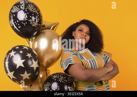 Porträt einer lächelnden jungen afroamerikanischen erwachsenen Frau, die auf gelbem Hintergrund süß aussieht und Ballons hält Stockfoto
