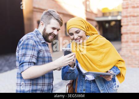 Schöner Mann zeigt ein Foto auf Smartphone junge arabische Frau. Schöne muslimische weibliche Studentin trägt leuchtend gelben Hijab kommuniziert mit Mann im Freien Stockfoto